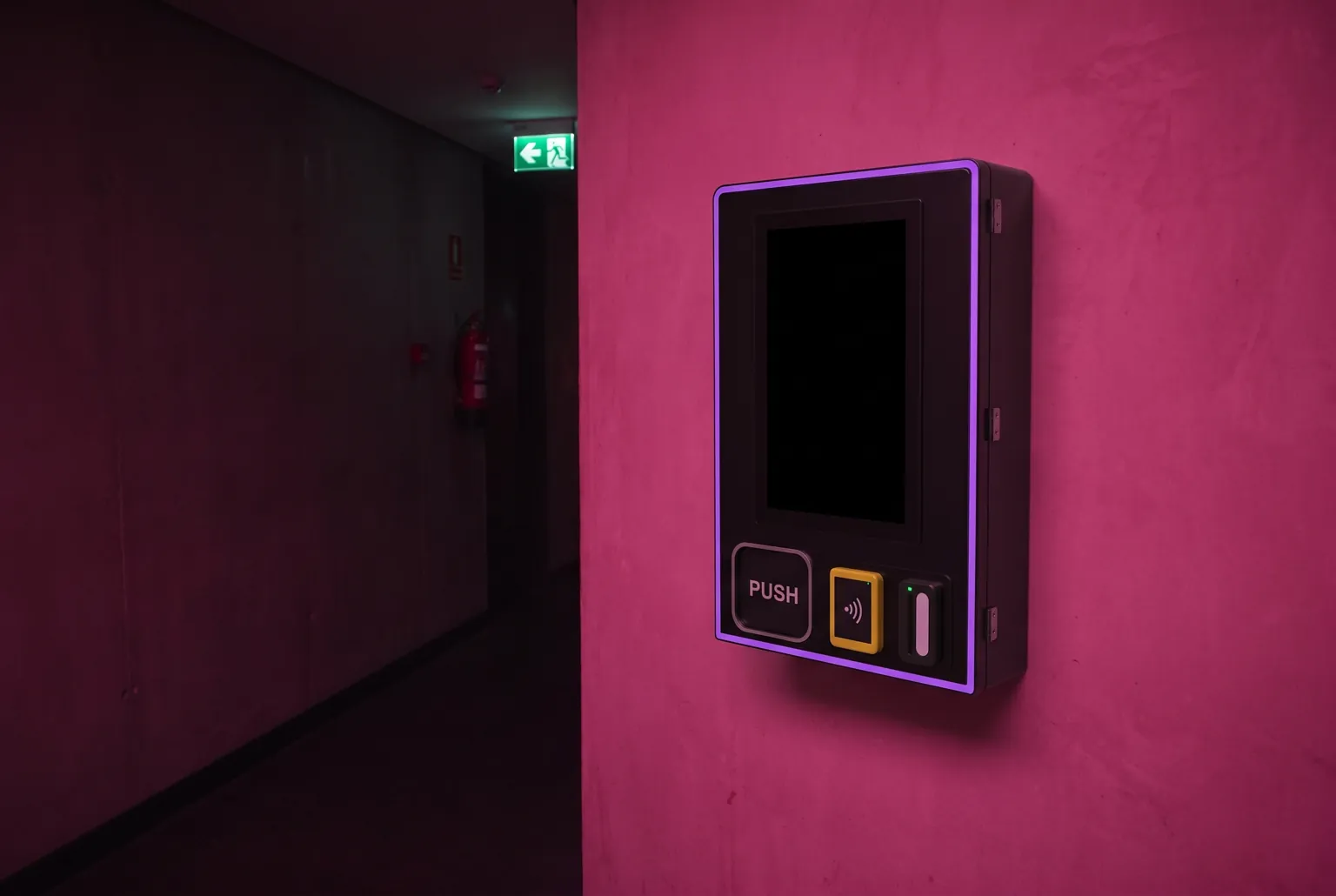 Le boîtier fixé au mur rose d’un couloir de club, à hauteur de poitrine, écran éteint. Au fond du couloir, dans la pénombre, un panneau de sortie de secours et un extincteur.
